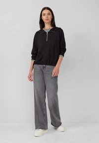 Felpa nera con zip e cappuccio, con dettagli a righe bianche sul colletto. Indossata con jeans larghi grigi chiari e sneakers bianche.