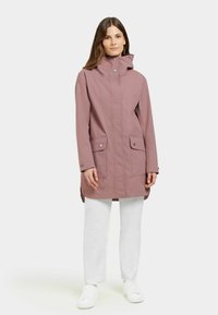 Rosa vattentät jacka med hög krage, två framfickor, tryckknappar och en avslappnad passform, bärs över vita byxor och sneakers.