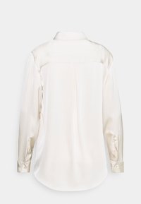 Blusa di seta color avorio con colletto classico, polsini abbottonati e una piega sul retro; texture liscia e orlo arrotondato.