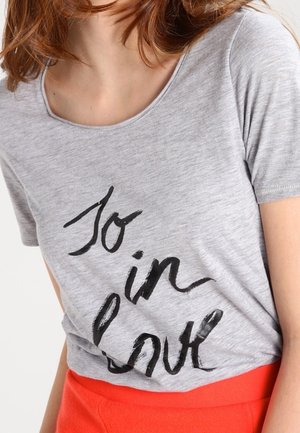 Kobieta w szarej koszulce z czarnym napisem "To in love" i czerwonej spódnicy, widoczna od szyi do bioder, na jednolitym tle.
