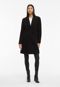 Cappotto di lana nero con colletto a picco, due bottoni e tasche laterali, abbinato a jeans neri e stivaletti, su uno sfondo neutro.