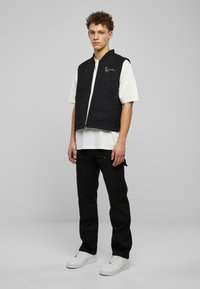 Melnā bezmuguras veste ar divām priekšējām kabatām un rāvējslēdzēju, valkāta virs baltā T-krekla. Melni bikses un baltas kedas papildina apģērbu.