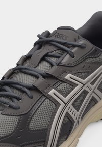 Κοντινό πλάνο γκρι και μαύρου αθλητικού παπουτσιού Asics με άνω μέρος από δίχτυ, λεπτομερή ραφή, αντανακλαστικές πλαϊνές ρίγες και μαύρα κορδόνια δεμένα σε φιόγκο.