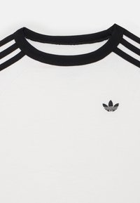 T-shirt de algodão branco com gola redonda canelada preta, três riscas pretas nos ombros e um pequeno logótipo preto bordado no peito.