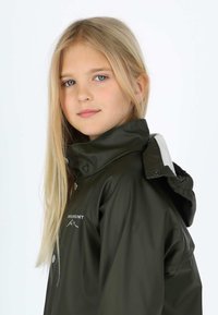 Jeune fille aux longs cheveux blonds portant une veste imperméable vert foncé avec un col montant et une capuche, légèrement de profil.