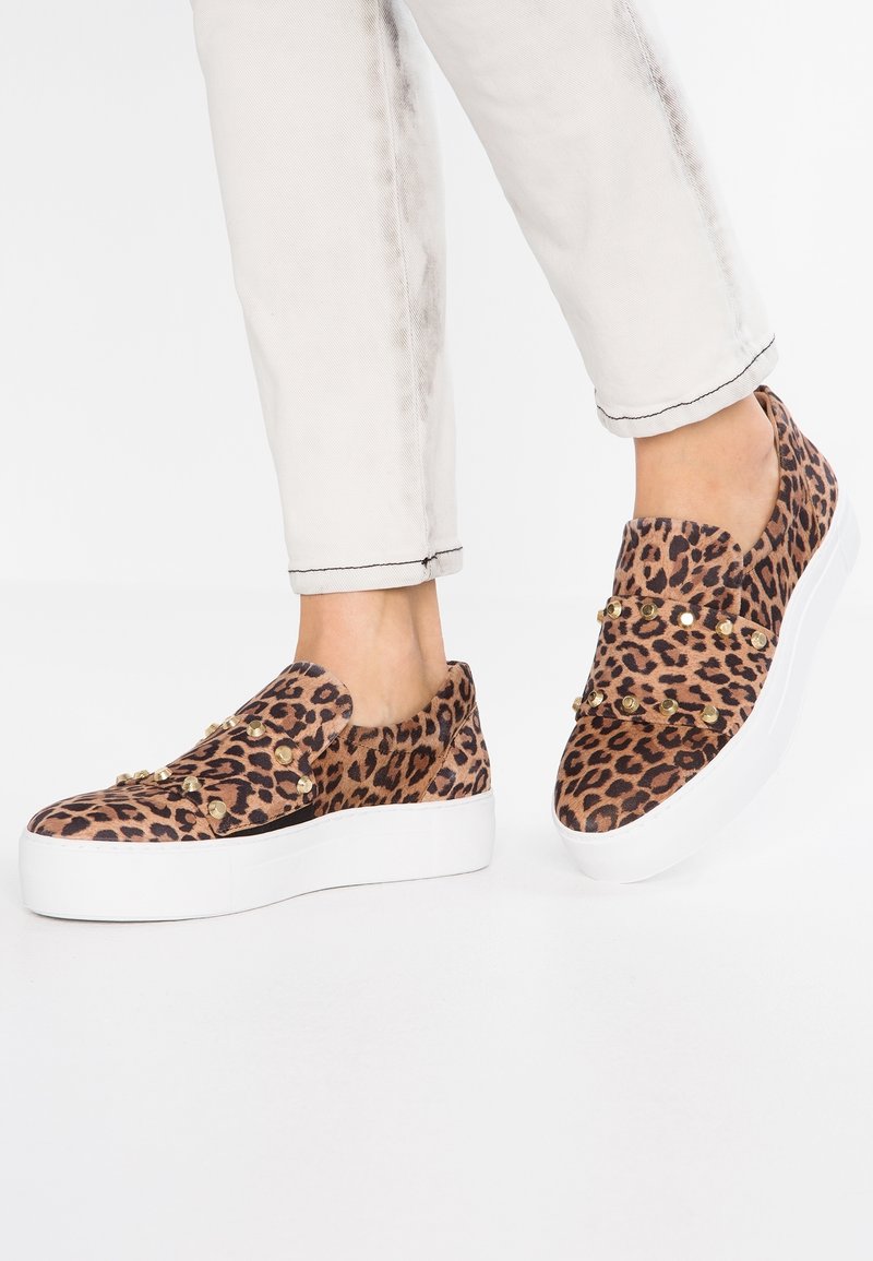 Schwarz-weiß gemusterte Slipper mit erhöhter weißer Sohle, verziert mit goldenen Nieten und einer weichen Textur, kombiniert mit hellen Jeans.