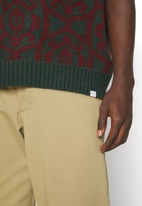 Stickad grön tröja med röd intrikat mönster, ribbad fåll, tillsammans med beiga byxor. Lappen synlig på tröjans nederkant.