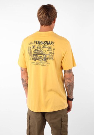 T-shirt en coton jaune avec un design graphique au dos, représentant un personnage de dessin animé dans un atelier d'artiste, avec des planches de surf en arrière-plan.