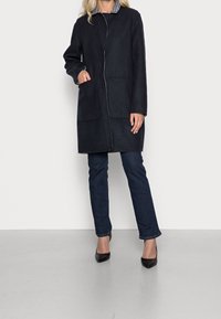 Donna che indossa un cappotto nero fino al ginocchio con grandi tasche frontali, jeans blu scuro e décolleté neri con punta, in piedi contro uno sfondo neutro.