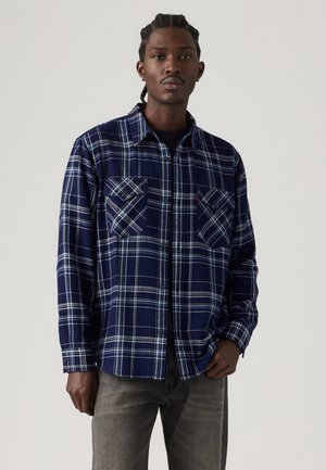 Homme aux cheveux tressés portant une chemise à manches longues à carreaux bleu marine et blanc et un pantalon gris, debout devant un fond clair uni.