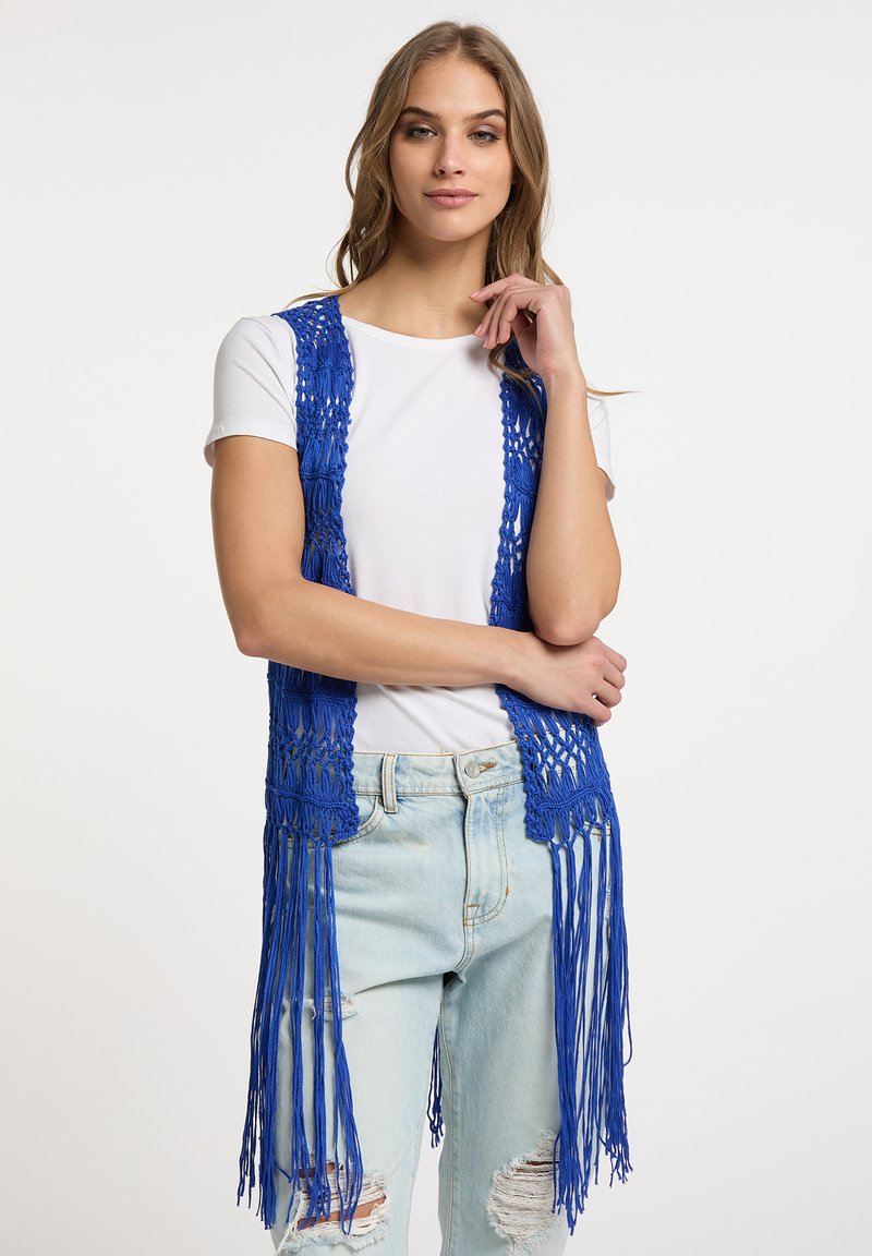 Blå virkad väst med öppet mönster och långa fransar, bärs över en vit t-shirt och ljustvättade slitna jeans.