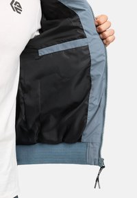 Chaqueta ligera con exterior azul, forro negro, puños acanalados y un bolsillo con cremallera. Presenta una cremallera completa y una etiqueta colgante.
