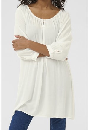 Femme aux cheveux bouclés portant un chemisier blanc ample à manches longues et un jean bleu, les bras croisés au niveau de la taille.