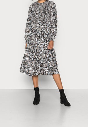 Woman wearing a long-sleeve, black floral midi dress with a smocked bodice, paired with black ankle boots, standing on a gray floor.