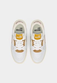 Άσπρα και μπεζ αθλητικά παπούτσια Love Moschino με λευκά κορδόνια και κίτρινες ετικέτες με το λογότυπο στις γλώσσες, από πάνω σε λευκό φόντο.