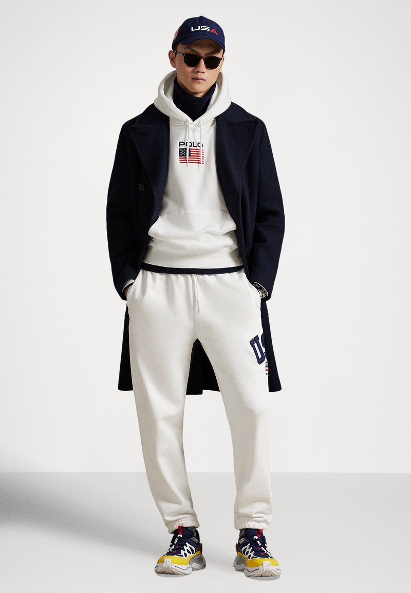 Manteau bleu marine sur un sweat à capuche blanc avec impression de drapeau, pantalon de survêtement crème avec logo "USA", lunettes de soleil, et baskets multicolores.
