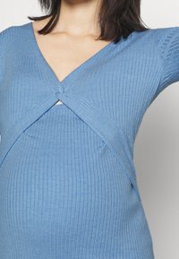 Femme portant un haut en maille côtelée bleu avec un col en V torsadé, coupé au milieu du torse, avec des cheveux noirs tombant sur les épaules.