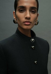 Black tailored jacket with a high collar, featuring large round buttons. Model has statement gold hoop earrings and a neutral makeup look.