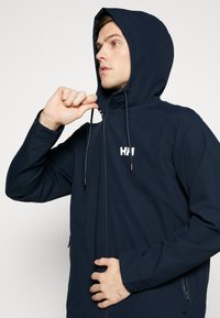 Chaqueta navy con cremallera y capucha, con un logo blanco, cordones ajustables y bolsillos laterales. Fabricada con un material ligero y suave.