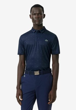 Uomo che indossa una polo blu navy a maniche corte con piccoli motivi, pantaloni blu navy e una cintura con fibbia metallica Lacoste.