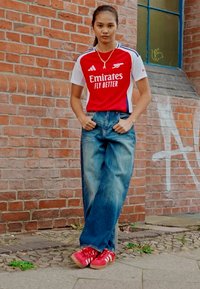 Červený a bílý sportovní dres s krátkým rukávem a textem "Emirates Fly Better", doplněný volnými modrými džínami a červenými teniskami. Cihlové pozadí.