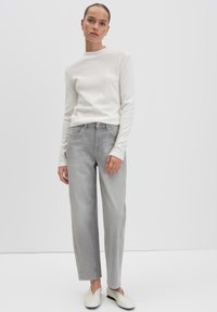 Graue, weite Jeans kombiniert mit einem enganliegenden weißen Langarmpullover und cremefarbenen Slippern, vor einem minimalistischem Hintergrund.