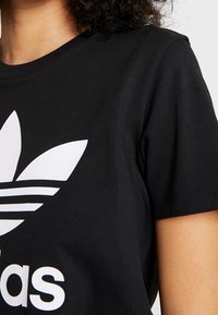 Černé tričko s krátkým rukávem, které má na přední straně velké bílé logo Adidas a klasický design tří pruhů. Jemná bavlněná textura.