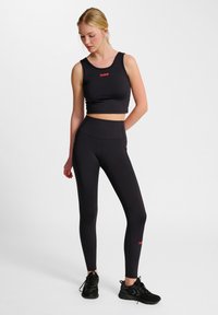 Svart sportig crop top med röd logotyp, kombinerad med högmidjade svarta leggings med texturerade sidopaneler. Svarta träningsskor fulländar looken.