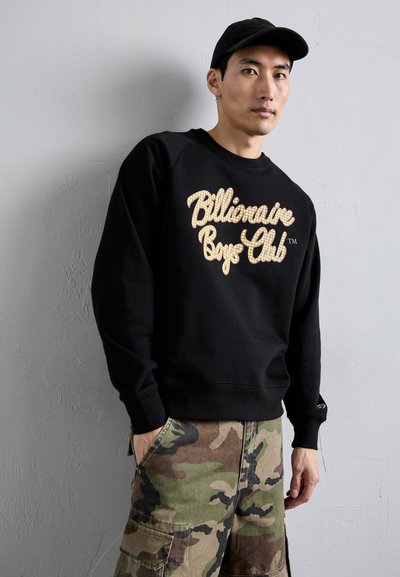 Melnā sporta jakā ar zeltītu, reljefu "Billionaire Boys Club" logo, kopā ar kamuflāžas krāsas cargo biksēm. Ikdienišķs dizains ar ribotiem apkakles galiem.