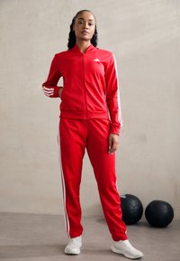 Červený atletický súprava z polyesteru s bielymi pruhmi pozdĺž rukávov a nohavíc. Bunda na zips a priliehavé nohavice, doplnené bielymi teniskami.