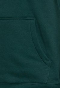 Tessuto di felpa verde scuro con una consistenza liscia, dotato di una tasca frontale e cuciture strette lungo l'orlo e i bordi della tasca.