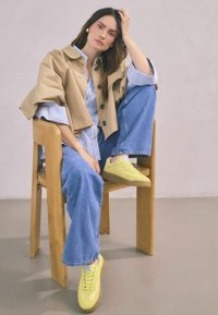 Veste beige oversize sur une chemise rayée bleue et blanche, associée à un jean en denim bleu clair et des baskets jaunes, assise sur une chaise en bois.