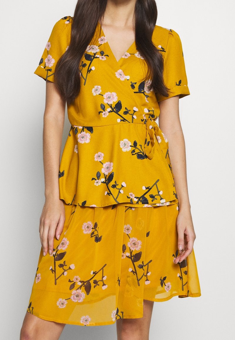 Femme portant une robe portefeuille fleurie jaune moutarde à manches courtes et jupe fluide, avec des motifs floraux roses et noirs.