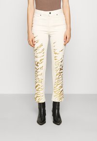 Jeans color crema con patrones abstractos dorados, con corte recto y cintura alta. Combinados con botas de tacón fino de punta negra.