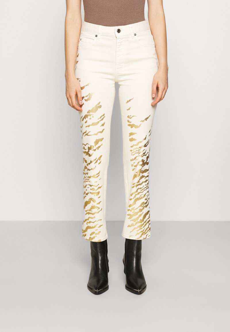 Jeans color crema con patrones abstractos dorados, con corte recto y cintura alta. Combinados con botas de tacón fino de punta negra.