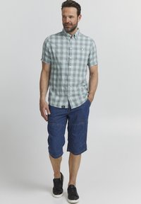 Camicetta a maniche corte a quadri verdi e bianchi con taschino, abbinata a pantaloni corti blu scuro fino al ginocchio e scarpe da ginnastica nere.