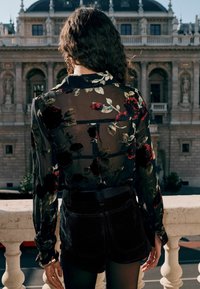 Schwarze transparente Bluse mit floralen Mustern, langen Ärmeln und hohem Kragen, kombiniert mit schwarzen hochgeschnittenen Shorts. Dekorative Beschläge sind sichtbar.