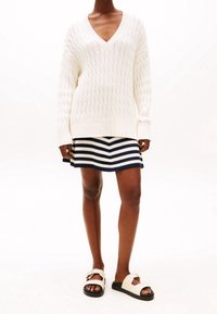 Bílý pletený svetr s výstřihem do V, spárovaný s námořnickou modrou sukní s bílými pruhy a světlými sandály s detaily přezky.