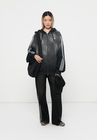 Svart zip-up hoodie med vita randdetaljer, ihop med mörka vida byxor och en svart keps. Slät textur, avslappnad sportig design.