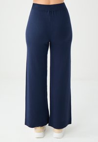 Marineblauwe wijde pijpen met een geribbelde tailleband en een gladde textuur, met een rechte snit en aansluitende enkelmanchetten. Gedragen met lichte sneakers.