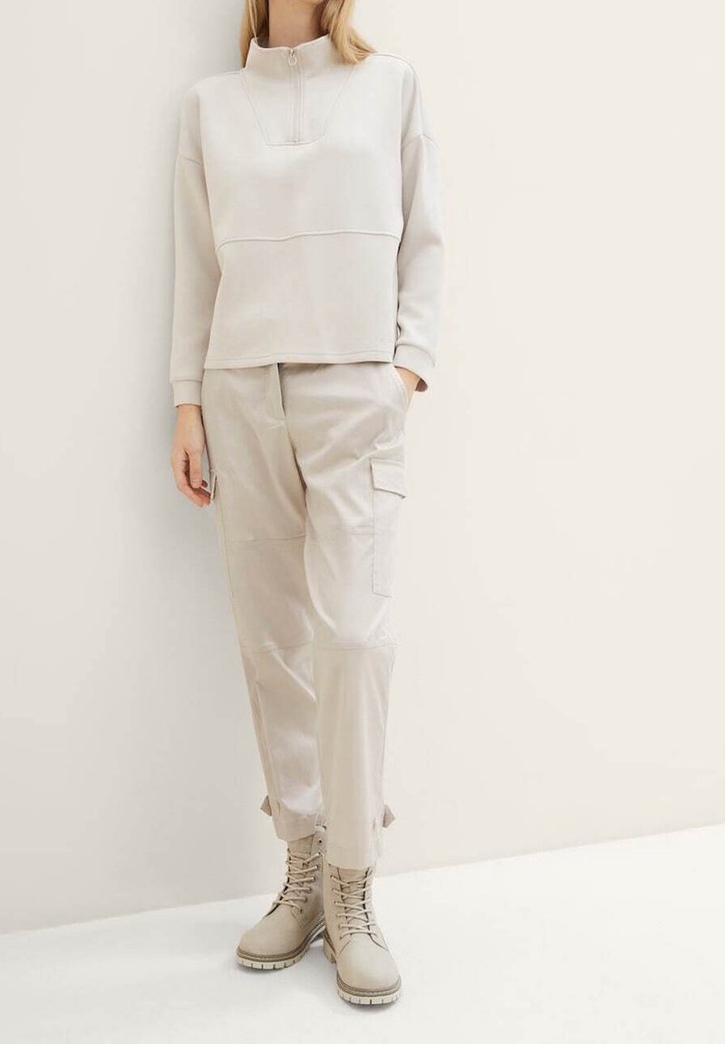 Femme portant un sweat à capuche beige à fermeture éclair, un pantalon cargo avec poches latérales et des bottines à lacets, debout contre un mur clair.
