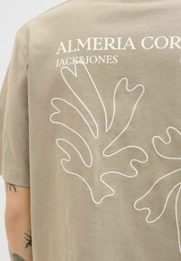Beige Baumwoll-T-Shirt mit einem großen weißen Blattdesign auf der Rückseite und dem Text "ALMERIA COR" darüber in fetter Schrift.