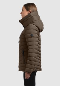 Bruine geïsoleerde jas met een puffy ontwerp, grote capuchon, zijzakken en een zwart logo-patch op de mouw. Gevoed, warm materiaal.