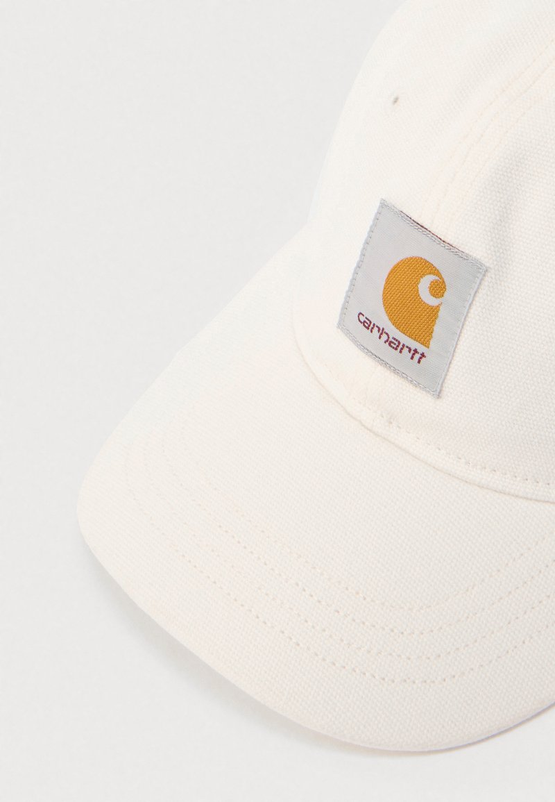 Cappellino in tessuto Carhartt color avorio con visiera curva e toppa logo quadrata cucita con emblema arancione e nome del marchio.