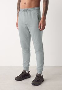 Pantaloni sport de culoare gri deschis, cu talie elastică, buzunare laterale cu fermoar și design conic al piciorului. Purtați cu pantofi sport închiși la culoare.