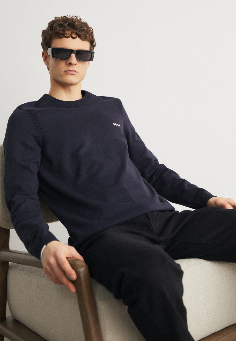 Mornarska sweatshirt s ovratnikom iz mehkega materiala z rebrastimi detajli, v kombinaciji z črnimi kavbojkami. Model nosi temne sončne očala in sedi.