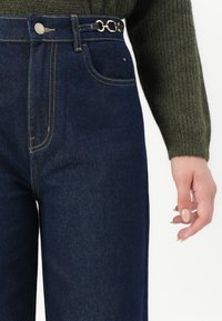 Jeans iz jeansa z visokim pasom v temno modri barvi z vidnimi šivi, s srebrnim verižnim poudarkom na zankah za pas in sprednjim žepom.