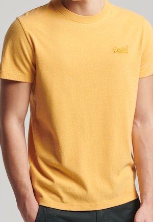 Homme portant un t-shirt jaune à manches courtes avec un petit logo brodé sur la poitrine gauche, les mains reposant dans les poches du pantalon.