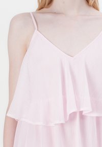 Rózsaszín, ujjatlan ruha V-kivágással és réteges fodros részlettel. Könnyű anyagból készült, sima, enyhén texturált felülettel.