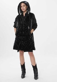 Czarna pikowana bezrękawnik z kapturem, błyszcząca powierzchnia, z przodu guziki i boczne kieszenie, noszony na czarnej sukience i zestawiony z czarnymi botkami.